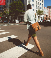 A person wearing a light shirt, shorts, and brown shoes carries the Bed|Stü Hue messenger bag—with its adjustable crossbody strap—as they cross a city street on a sunny day, effortlessly blending work-to-weekend style.