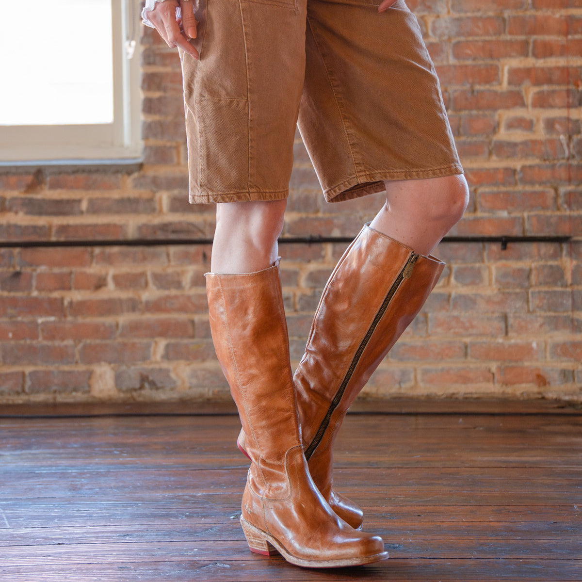 A person wearing the Humble knee-high, square-toe brown leather boots with a wooden heel by Bed|Stü, paired with tan knee-length shorts, stands on a wooden floor in front of a brick wall.