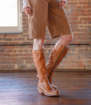 A person wearing the Humble knee-high, square-toe brown leather boots with a wooden heel by Bed|Stü, paired with tan knee-length shorts, stands on a wooden floor in front of a brick wall.