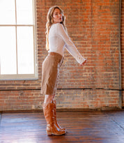 A woman stands sideways on a wooden floor in front of a brick wall, wearing a white long-sleeve top, tan knee-length skirt, and the "Humble" tall leather boots with a wooden heel by Bed|Stü.