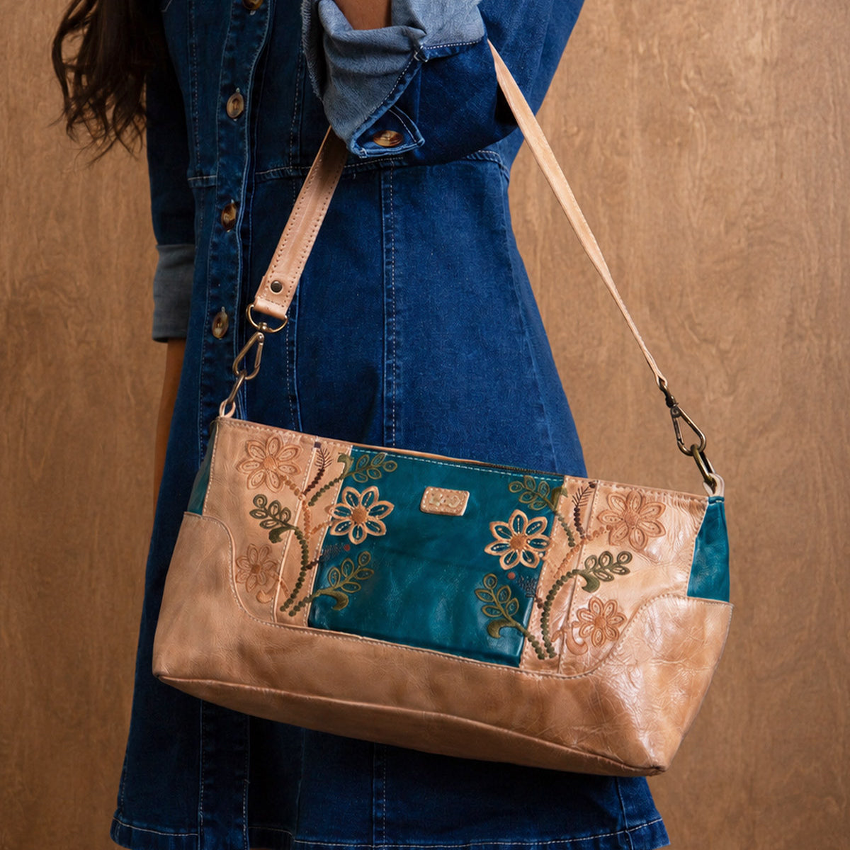A person wearing a blue denim dress holds the Bed|Stü Hyperbolic handcrafted leather shoulder bag, featuring floral embroidery and bohemian flair, against a brown background.