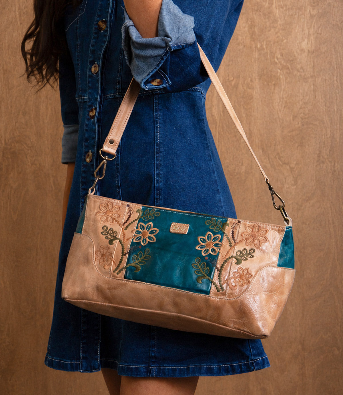 A person wearing a blue denim dress holds the Bed|Stü Hyperbolic handcrafted leather shoulder bag, featuring floral embroidery and bohemian flair, against a brown background.