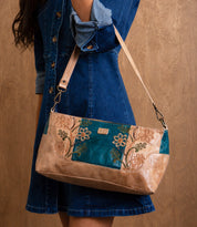 A person wearing a blue denim dress holds the Bed|Stü Hyperbolic handcrafted leather shoulder bag, featuring floral embroidery and bohemian flair, against a brown background.