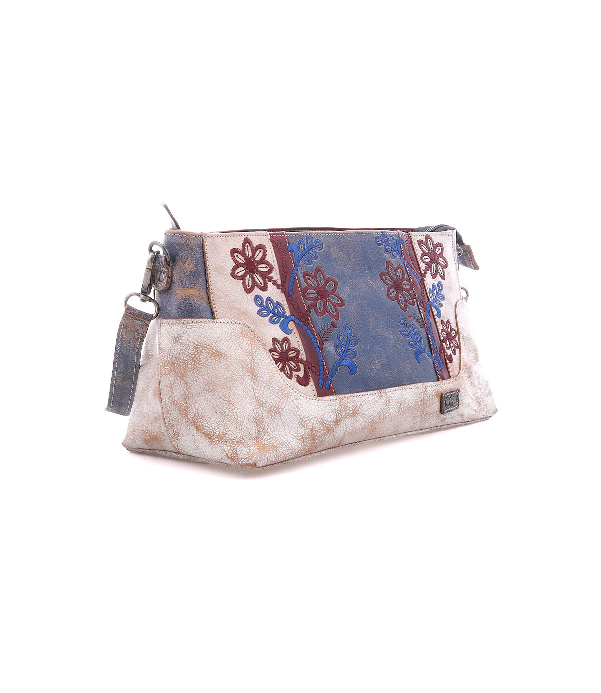 The Hyperbolic clutch by Bed|Stü is a handcrafted, bohemian-style beige accessory featuring a blue and maroon floral embroidered panel, zipper closure, and a detachable wrist strap, shown against a white background.