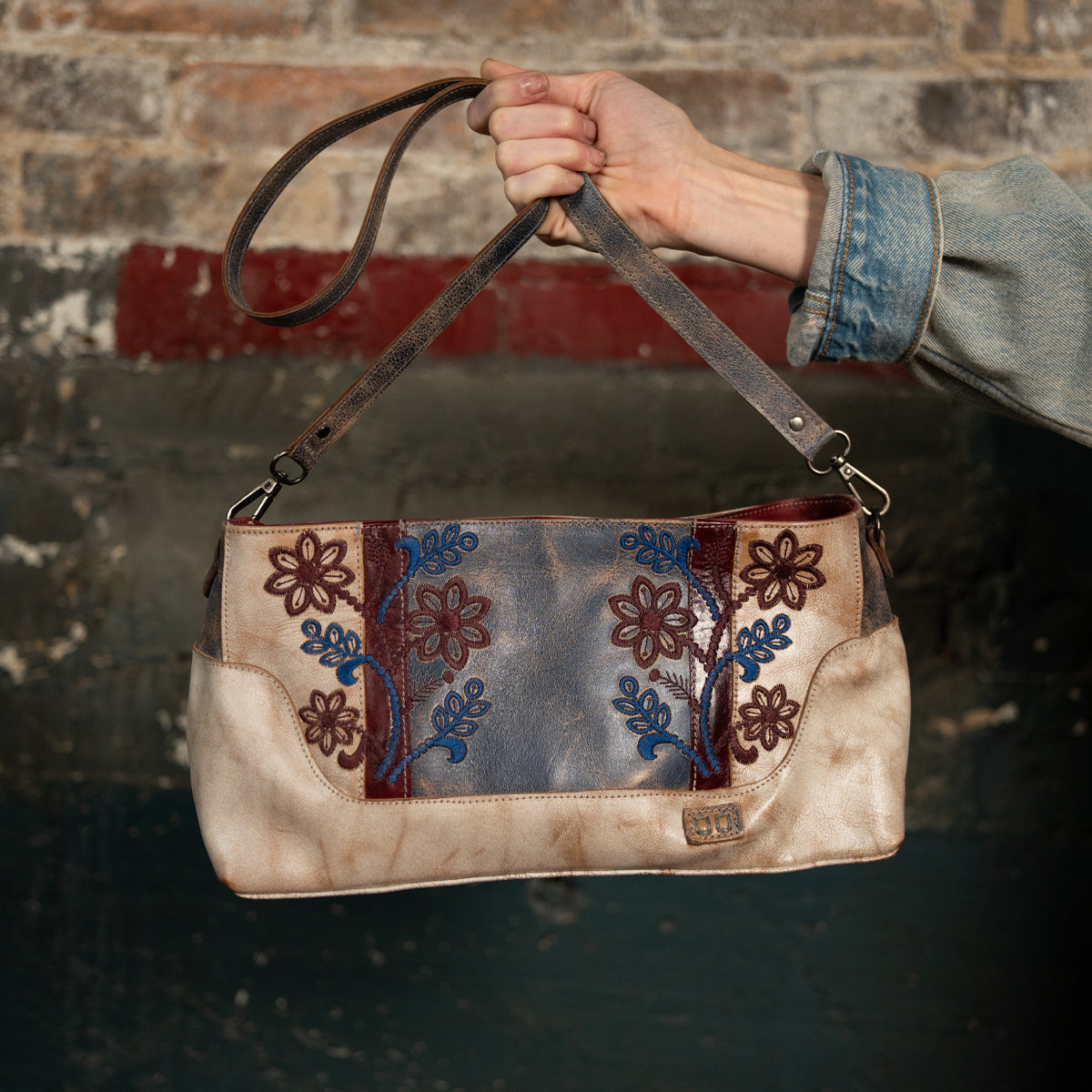 A hand holds the Bed|Stü Hyperbolic handcrafted leather shoulder bag, featuring blue and red floral embroidery, set against a weathered brick and painted wall backdrop, infusing the scene with bohemian charm.