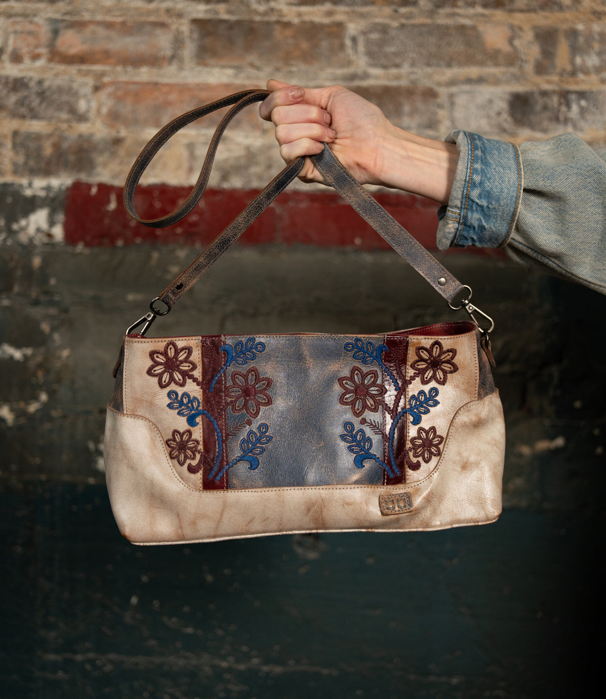 A hand holds the Bed|Stü Hyperbolic handcrafted leather shoulder bag, featuring blue and red floral embroidery, set against a weathered brick and painted wall backdrop, infusing the scene with bohemian charm.