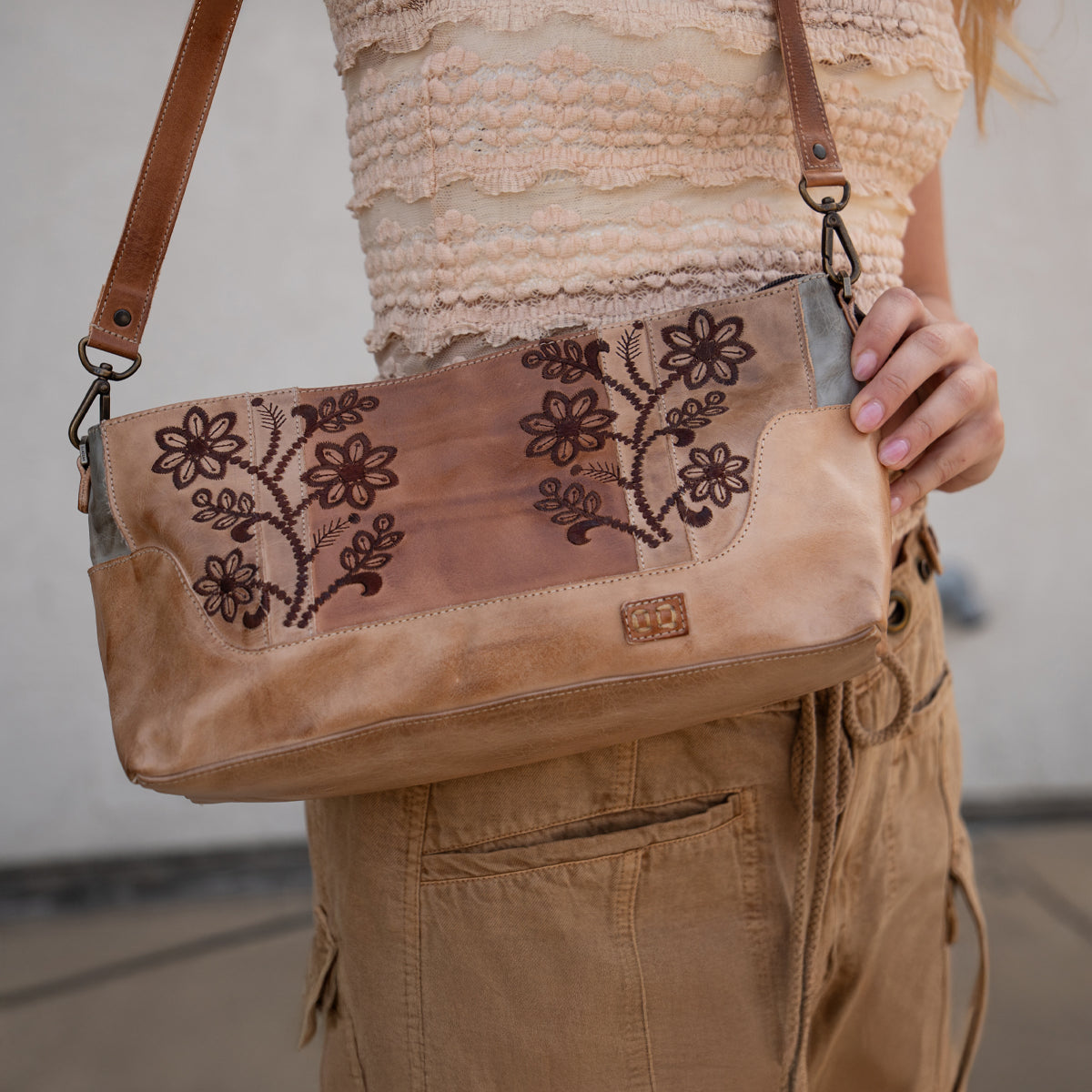 A person wearing a textured beige top and tan pants holds the Bed|Stü Hyperbolic bag—a bohemian leather shoulder bag featuring floral embroidery, a brown strap, and an effortlessly handcrafted charm.