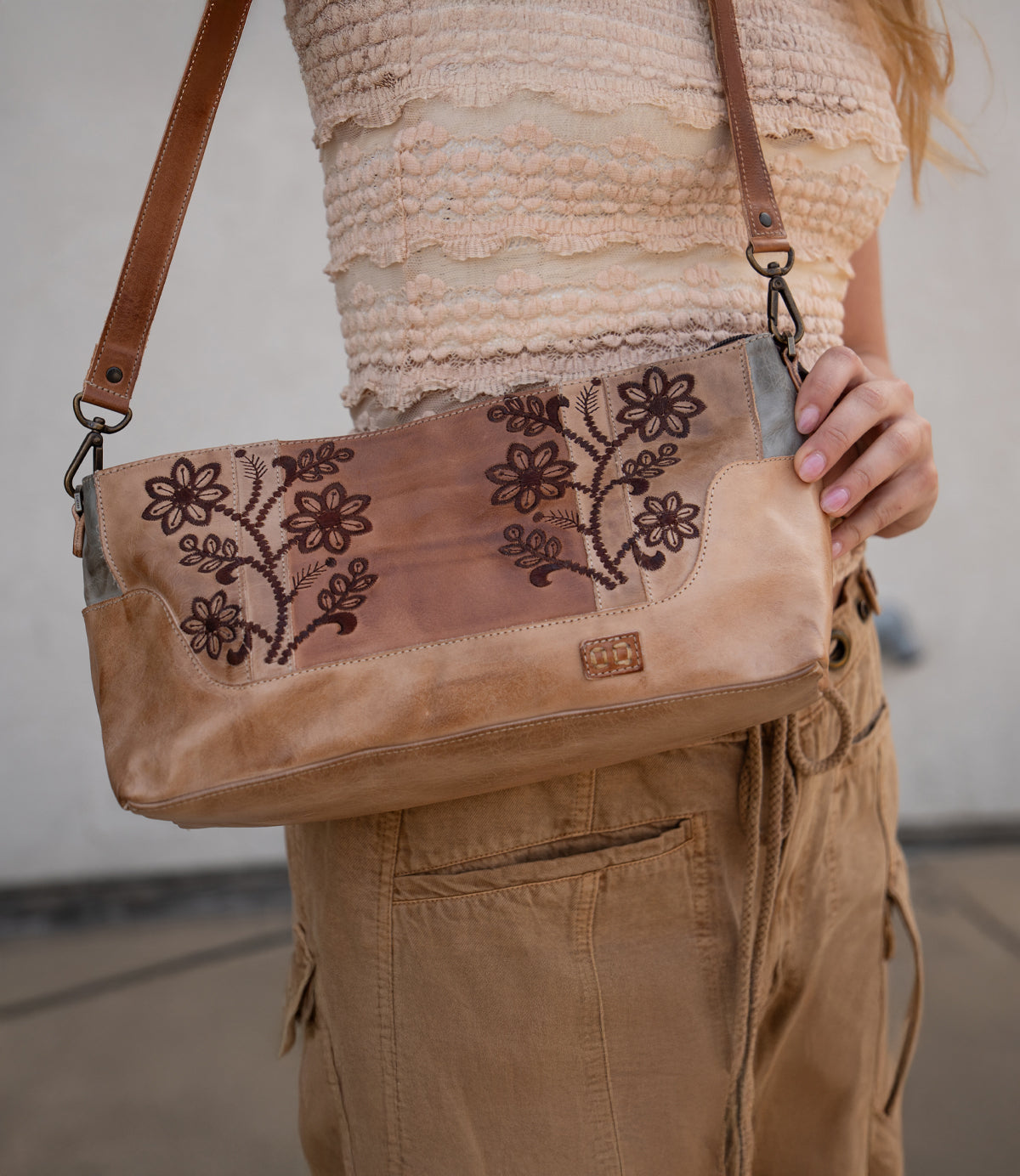 A person wearing a textured beige top and tan pants holds the Bed|Stü Hyperbolic bag—a bohemian leather shoulder bag featuring floral embroidery, a brown strap, and an effortlessly handcrafted charm.