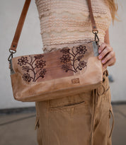 A person wearing a textured beige top and tan pants holds the Bed|Stü Hyperbolic bag—a bohemian leather shoulder bag featuring floral embroidery, a brown strap, and an effortlessly handcrafted charm.