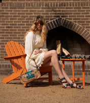 A woman in a white dress and sandals sits on an orange Adirondack chair near a fireplace, holding the Bed|Stü Hyperbolic handcrafted brown and green leather shoulder bag, wearing sunglasses and radiating effortless bohemian style.