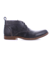 The Bed|Stü Illiad chukka boot, shown in profile on a white background, features black leather, a comfortable outsole with a brown sole, three eyelets for lacing, and a pull tab at the heel. This modern lace-up design blends classic style with everyday comfort.