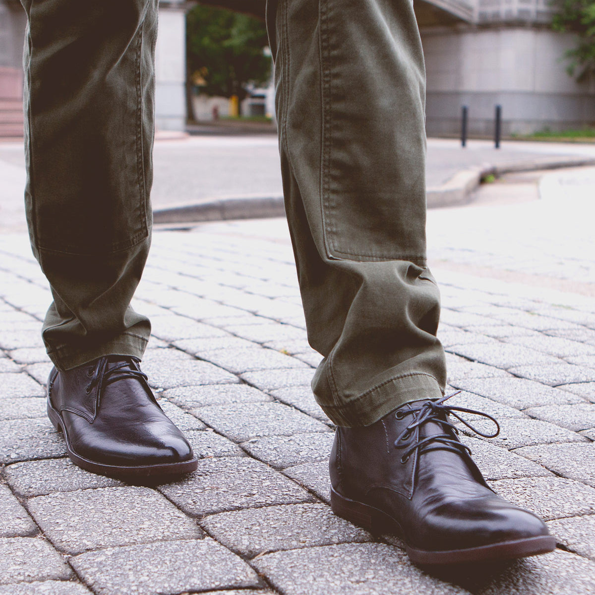 A person wearing olive green pants and Bed|Stü Illiad black leather chukka shoes with a comfortable outsole stands on a cobblestone sidewalk in an urban setting.