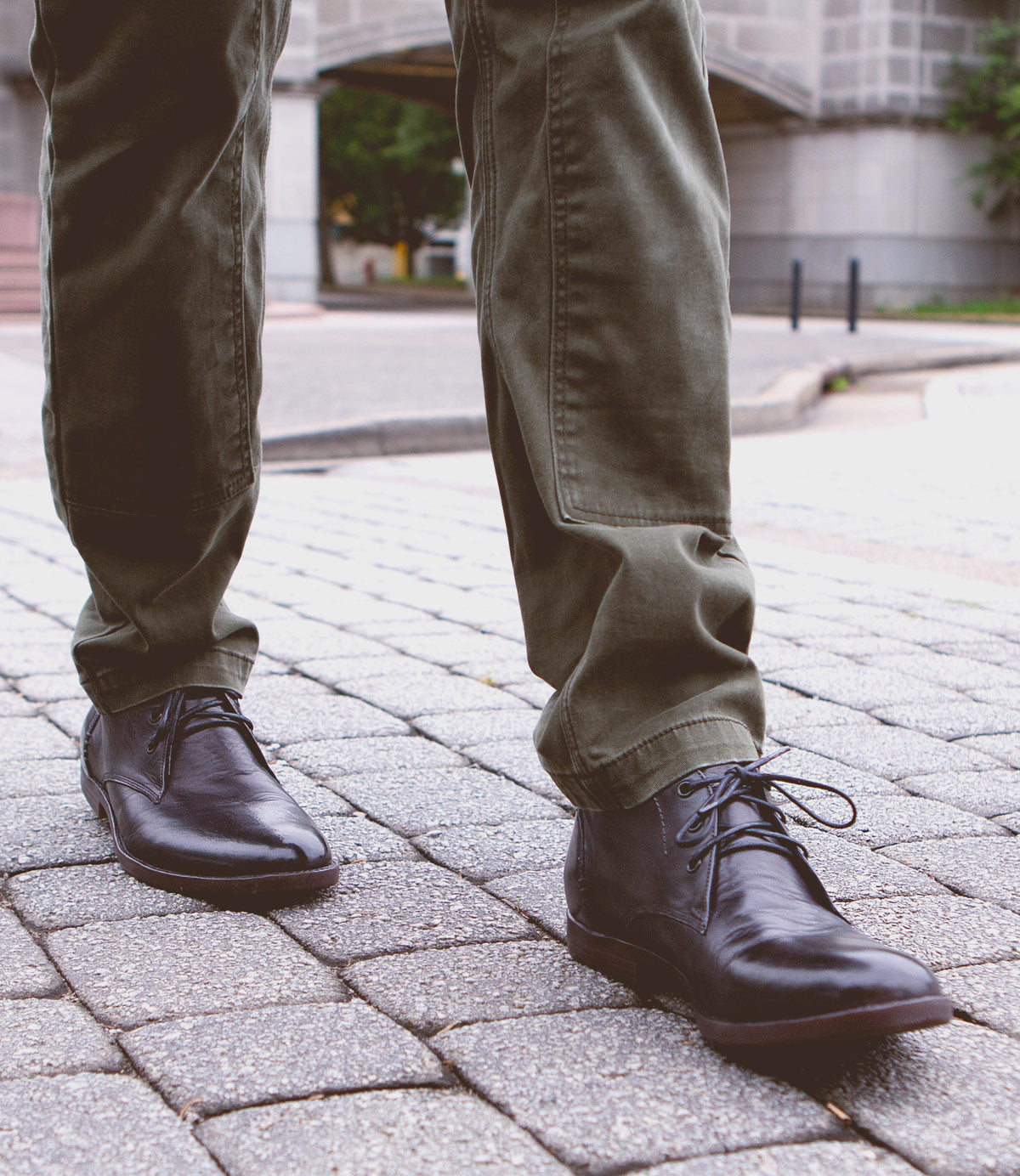 A person wearing olive green pants and Bed|Stü Illiad black leather chukka shoes with a comfortable outsole stands on a cobblestone sidewalk in an urban setting.