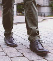 A person wearing olive green pants and Bed|Stü Illiad black leather chukka shoes with a comfortable outsole stands on a cobblestone sidewalk in an urban setting.