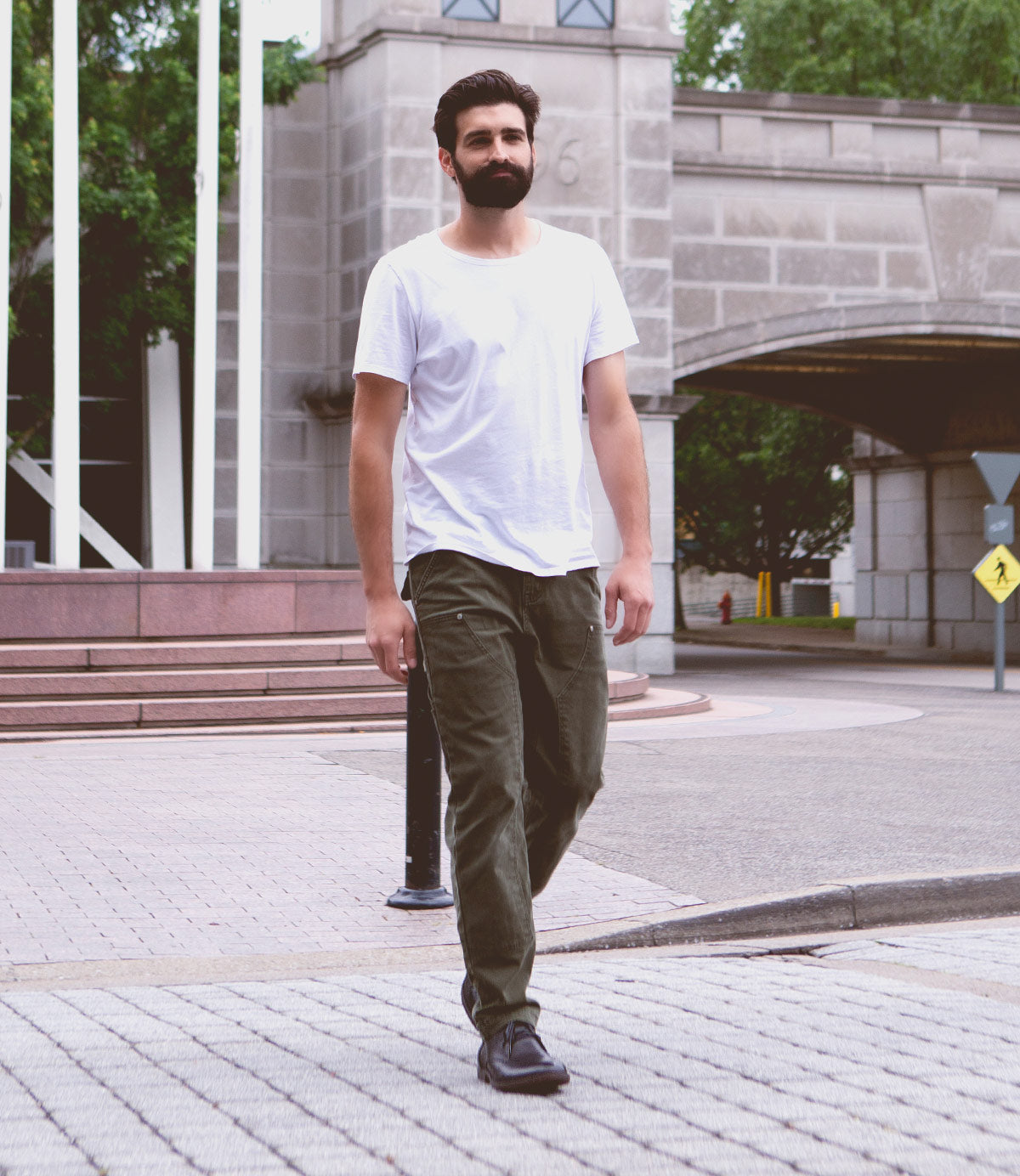 A bearded man walks on a paved street, wearing a white t-shirt, green pants, and the Illiad sleek black leather chukka shoes by Bed|Stü, featuring a comfortable outsole. A stone building and a pedestrian crossing sign appear in the background.