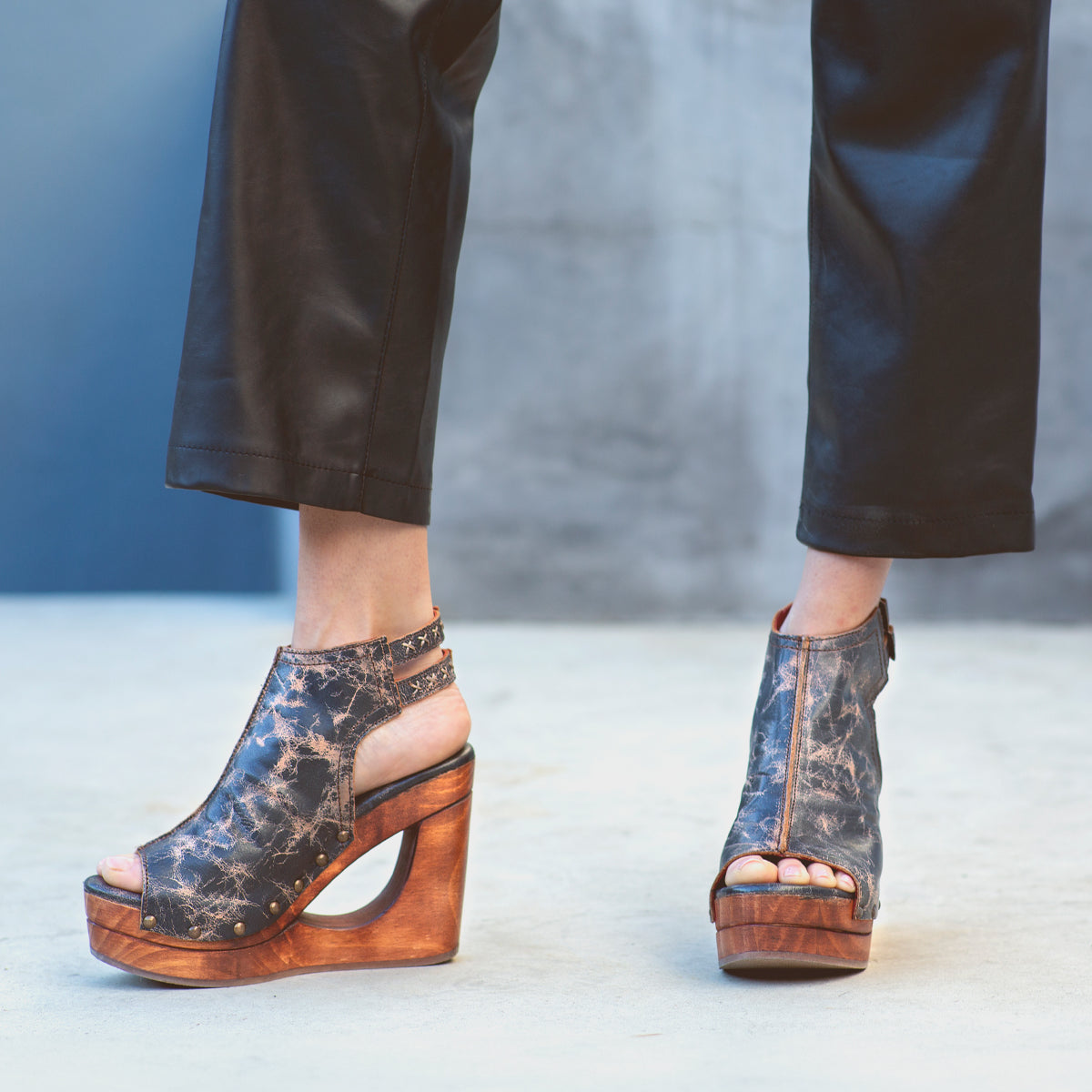 Wearing Bed|Stü’s Imelda black leather pants and wedge-heeled open-toe shoes featuring a bold cutout wedge and adjustable ankle buckles, the person stands on a light gray surface.
