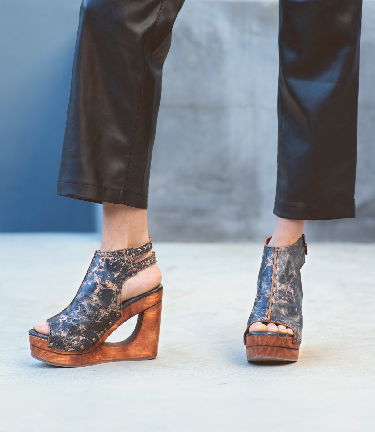 Wearing Bed|Stü’s Imelda black leather pants and wedge-heeled open-toe shoes featuring a bold cutout wedge and adjustable ankle buckles, the person stands on a light gray surface.