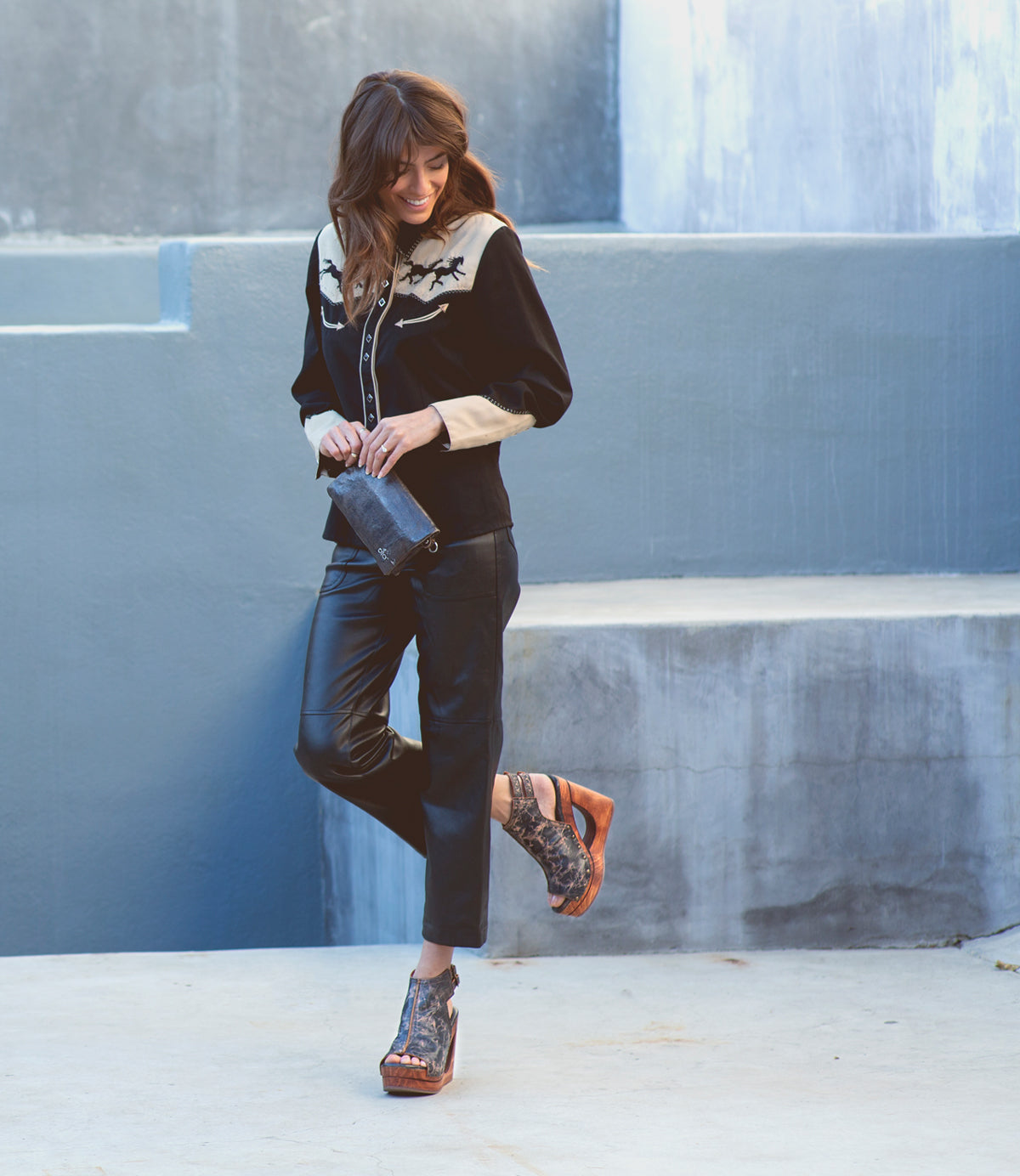 A woman stands outdoors against a concrete wall, wearing a black western-style shirt, black pants, and the Imelda cutout wedge sandals by Bed|Stü, featuring adjustable ankle buckles. Holding a small black bag, she looks down and smiles.
