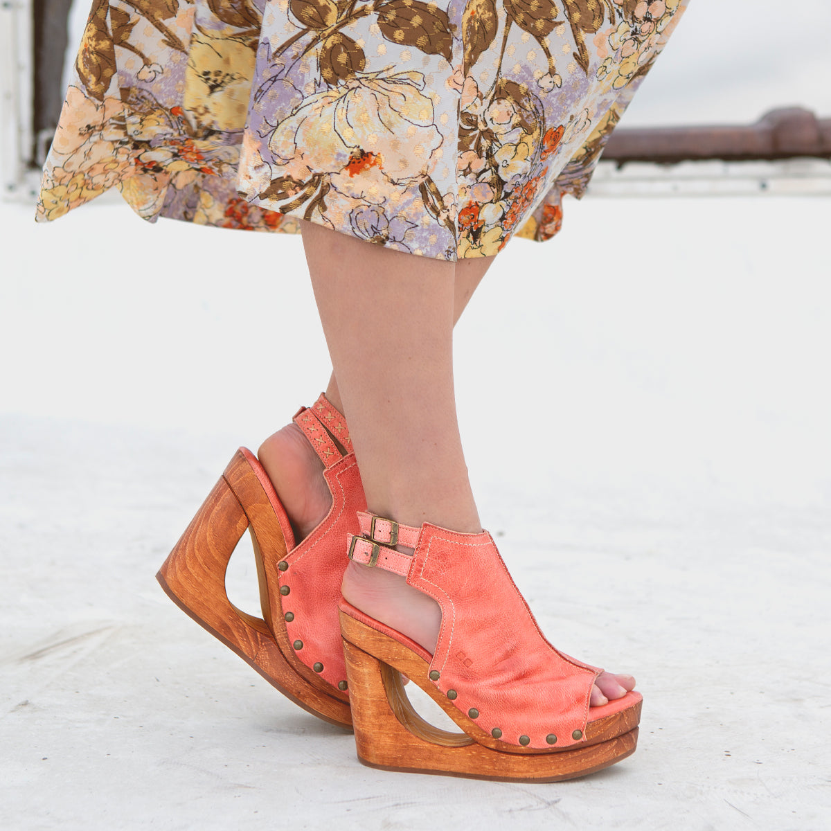 A person wearing a floral skirt and Bed|Stü Imelda coral peep-toe wedge sandals with wooden heels and an open-toe silhouette stands on a white surface.