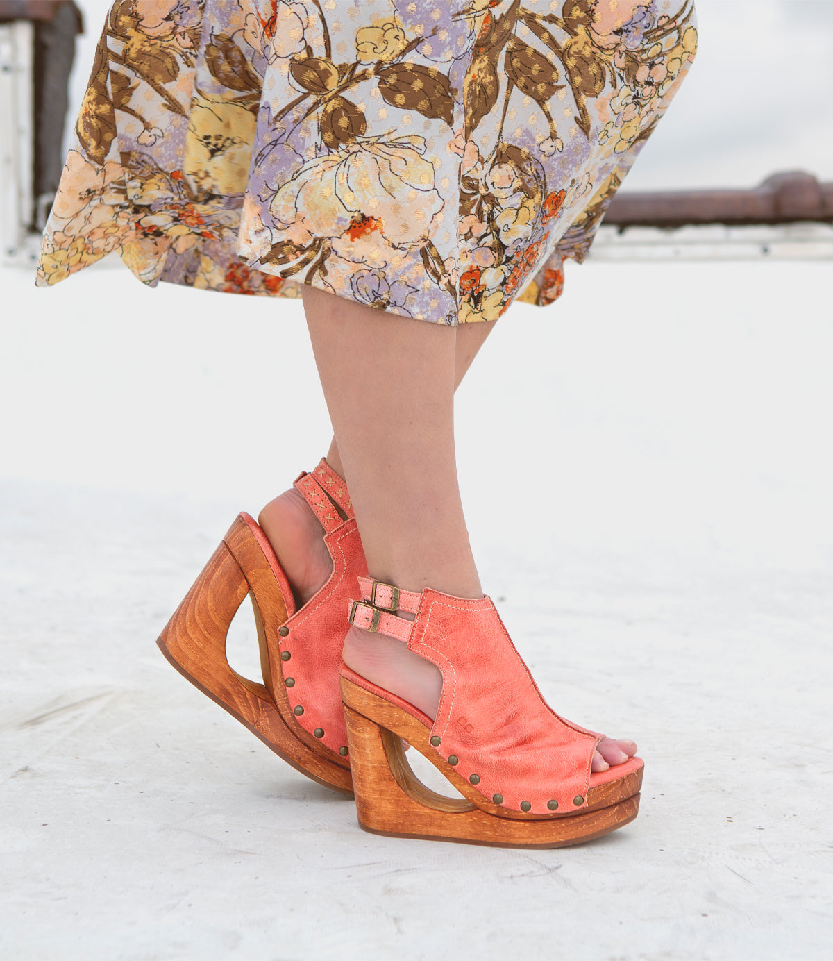A person wearing a floral skirt and Bed|Stü Imelda coral peep-toe wedge sandals with wooden heels and an open-toe silhouette stands on a white surface.