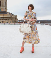 A woman in a floral dress and Bed|Stü Imelda coral platform shoes with an open-toe silhouette stands on a rooftop, holding a large white bag, with buildings and a cloudy sky in the background.