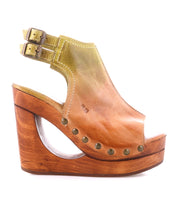 The Bed|Stü Imelda is a brown leather open-toe wedge sandal, showcased in a side view, featuring a stylish cut-out wooden heel, two adjustable ankle buckles, decorative brass rivets, and distinctive cutout wedge detailing.