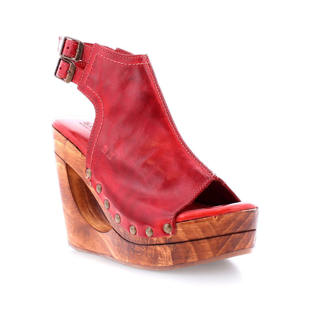 The Bed|Stü Imelda is a red leather open-toe wedge sandal featuring a bold cutout wedge design, wooden platform sole, metal stud accents, and an ankle strap with two adjustable buckles, shown against a white background.