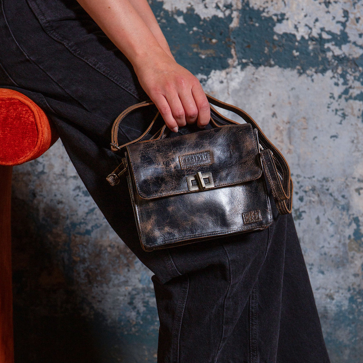 Seated on an orange chair against a textured wall, this individual wears black jeans and carries the Bed|Stü Improv—a small, vintage-inspired black leather mini purse featuring a buckle clasp, crossbody strap, and top handle.