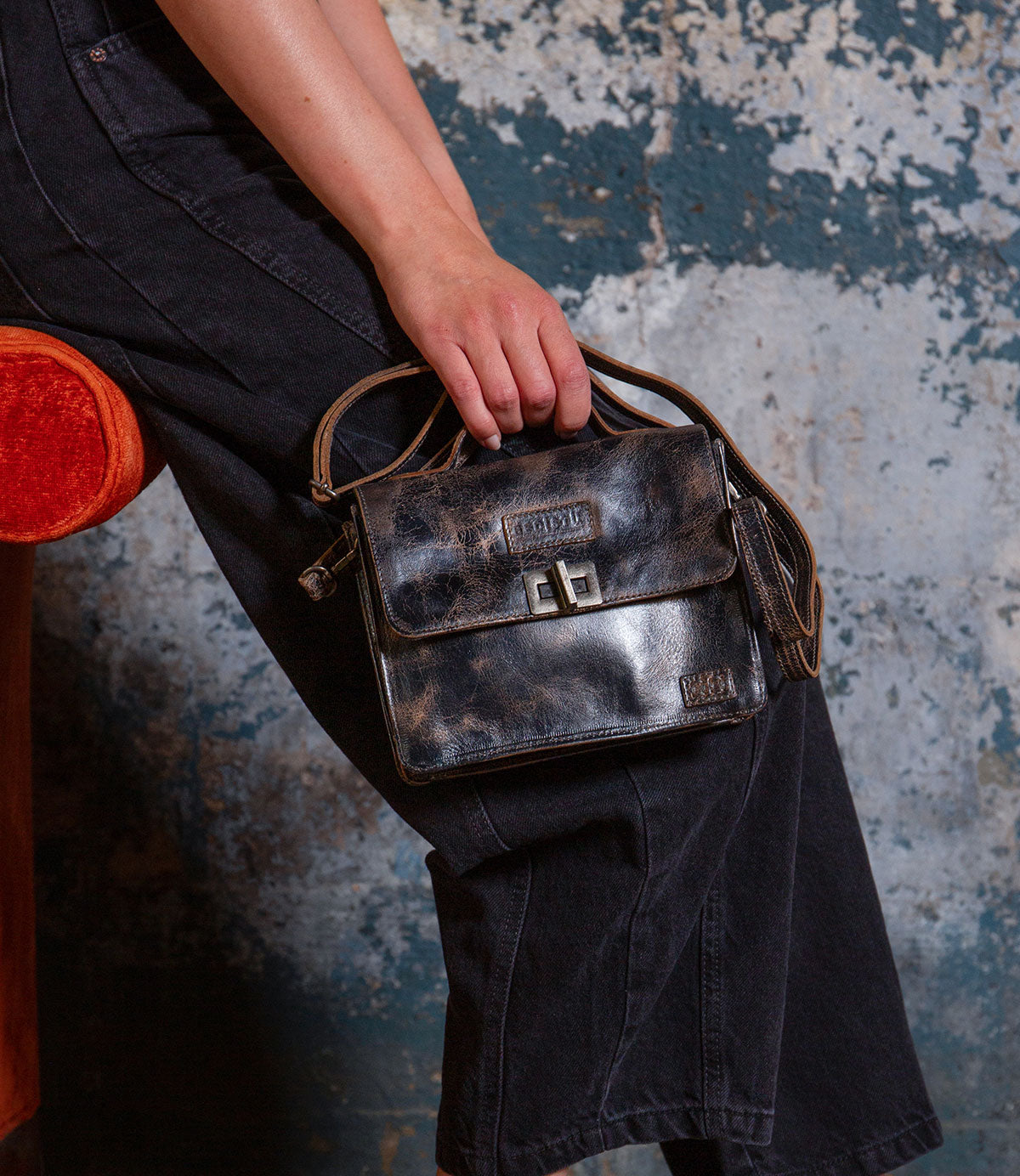 Seated on an orange chair against a textured wall, this individual wears black jeans and carries the Bed|Stü Improv—a small, vintage-inspired black leather mini purse featuring a buckle clasp, crossbody strap, and top handle.