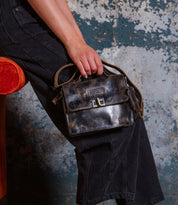 Seated on an orange chair against a textured wall, this individual wears black jeans and carries the Bed|Stü Improv—a small, vintage-inspired black leather mini purse featuring a buckle clasp, crossbody strap, and top handle.