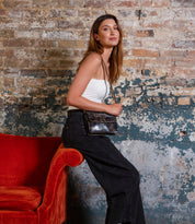 A woman in a white tank top and black pants sits on the arm of a red chair, holding the Bed|Stü Improv black mini purse with a top handle, in front of a distressed brick wall.