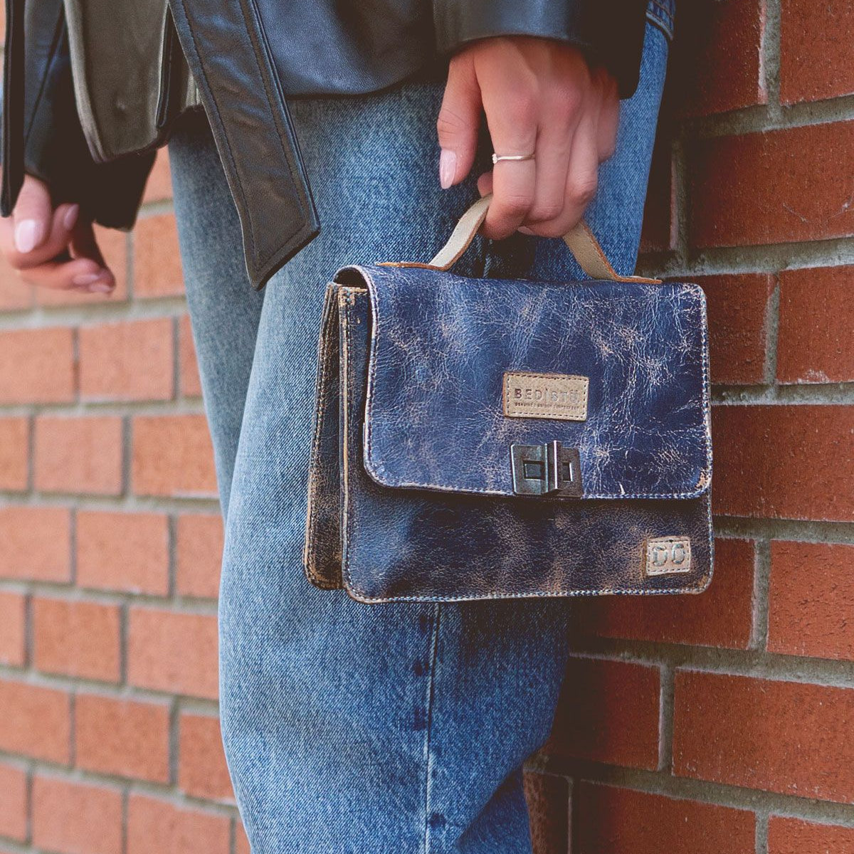 A person wearing blue jeans and a black leather jacket holds the Bed|Stü Improv, a small distressed navy leather satchel with a top handle, while standing next to a red brick wall.