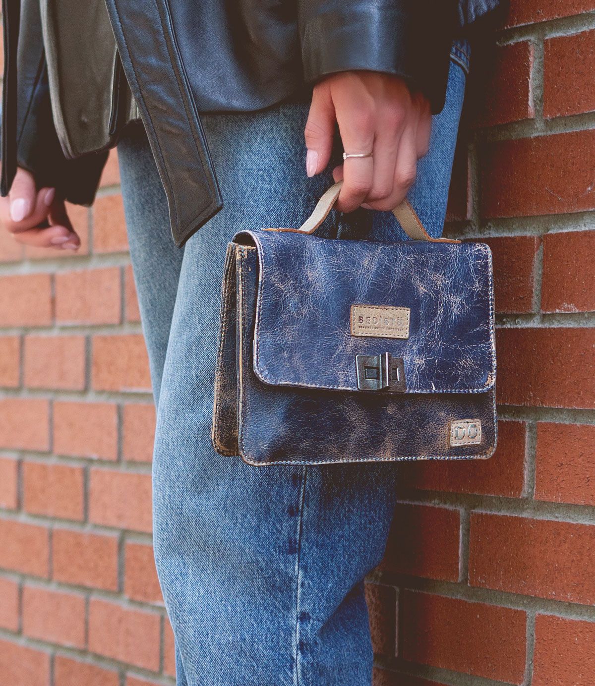 A person wearing blue jeans and a black leather jacket holds the Bed|Stü Improv, a small distressed navy leather satchel with a top handle, while standing next to a red brick wall.