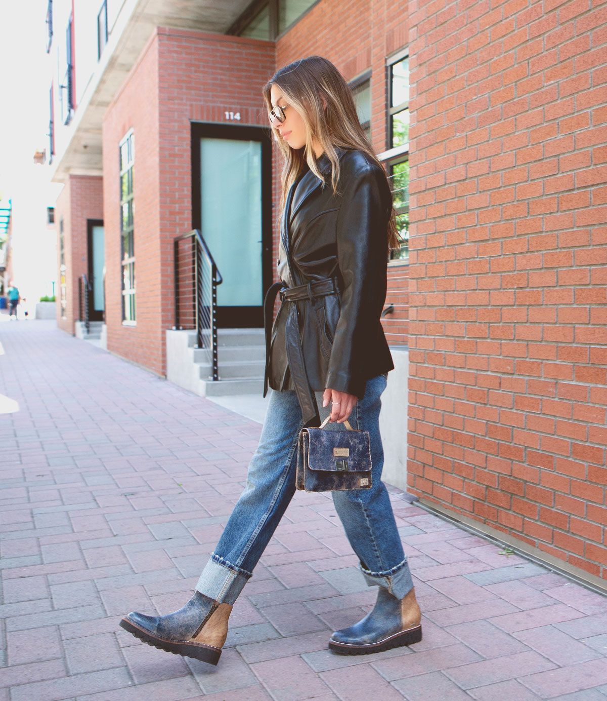 A woman walks on a city sidewalk wearing sunglasses, a black jacket, blue cuffed jeans, ankle boots, and carrying the Improv mini purse with a top handle by Bed|Stü.