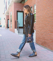 A woman walks on a city sidewalk wearing sunglasses, a black jacket, blue cuffed jeans, ankle boots, and carrying the Improv mini purse with a top handle by Bed|Stü.