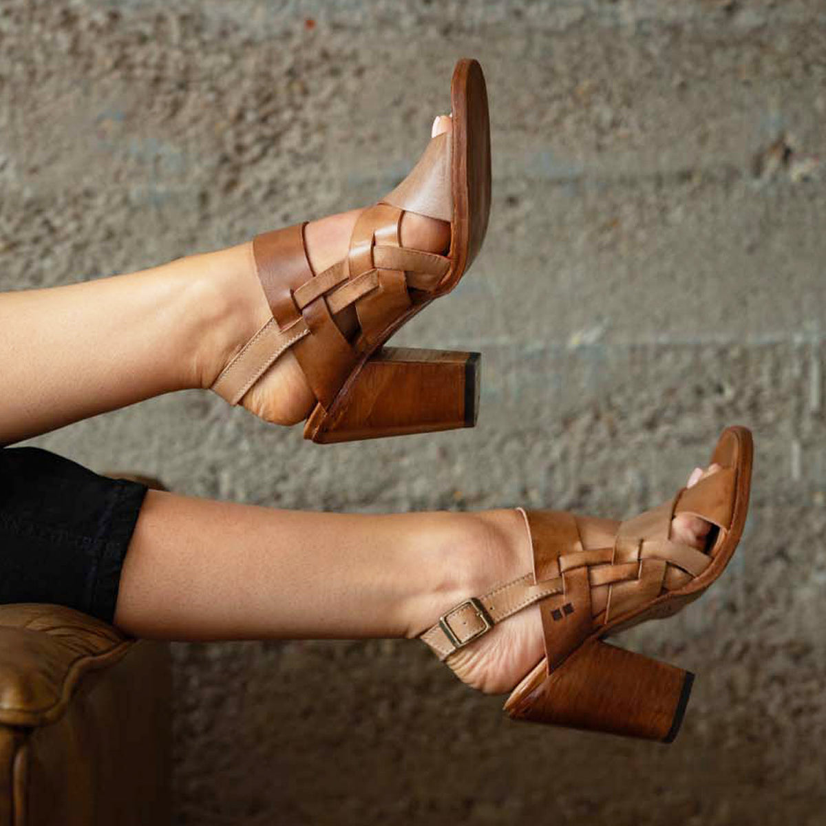 A person wears the Bed|Stü Intersection tan woven leather heeled sandals, featuring an open-toe design and adjustable ankle strap, while resting their feet on a brown chair against a textured gray wall.