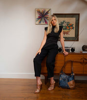 A woman in a black outfit and the Bed|Stü Intersection heels with an adjustable ankle strap leans against a wooden credenza with a radio, bag, and framed artwork behind her on a hardwood floor.