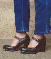 A person wearing blue jeans stands on a stone surface, showcasing the Bed|Stü Ishtar black Mary Jane pump high heel shoes with an adjustable strap.