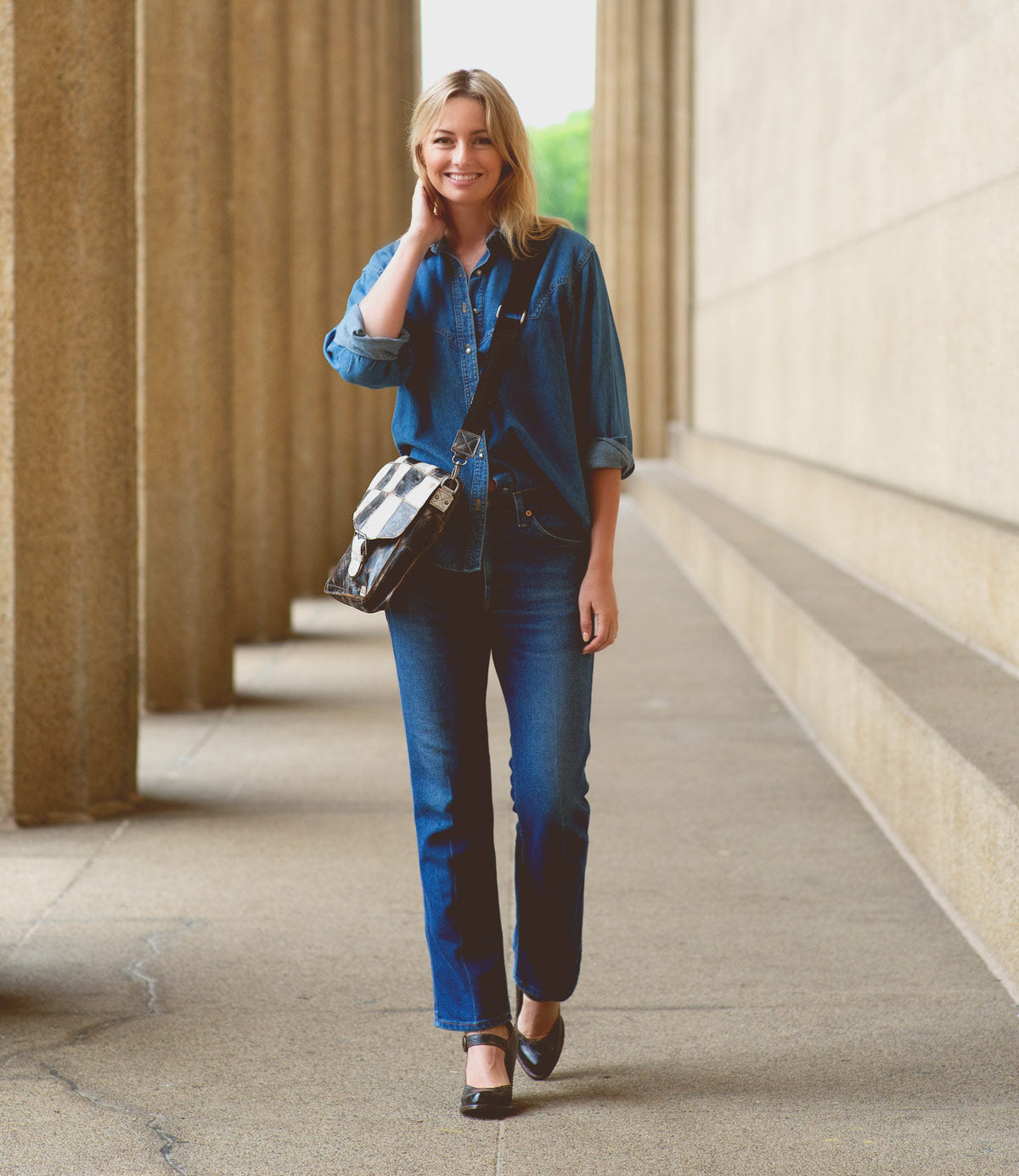 A woman in a blue denim shirt and jeans stands outdoors in a colonnade, smiling, with a checkered bag across her shoulder and stylish Bed|Stü Ishtar block heel Mary Jane pumps completing her look.