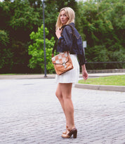 A woman stands on a cobblestone path, wearing a white dress, dark jacket, and Bed|Stü Ishtar brown Mary Jane pumps featuring an adjustable strap and block heel. She carries a tan shoulder bag, while trees and greenery are visible in the background.
