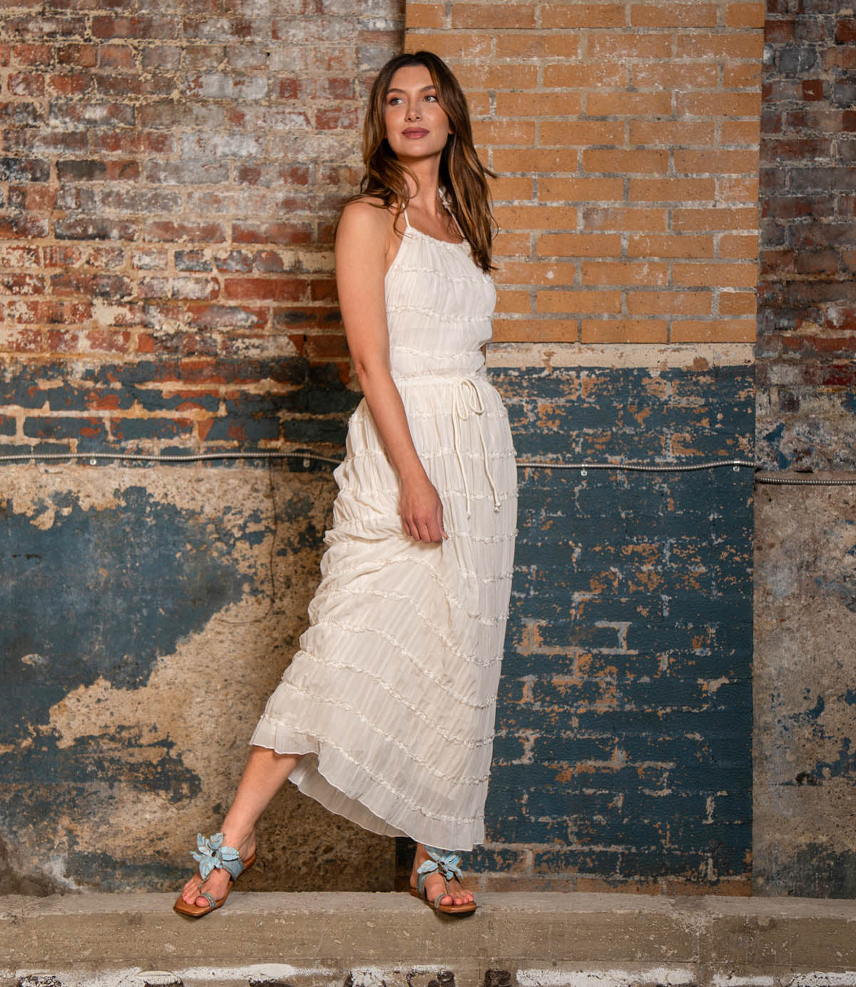 A woman in a sleeveless white dress stands against a textured brick and painted wall, one foot stepping forward as she gazes slightly to her left—her outfit completed by the Isobel leather sandal from Bed|Stü, featuring a braided strap that’s perfect for effortless summer style.