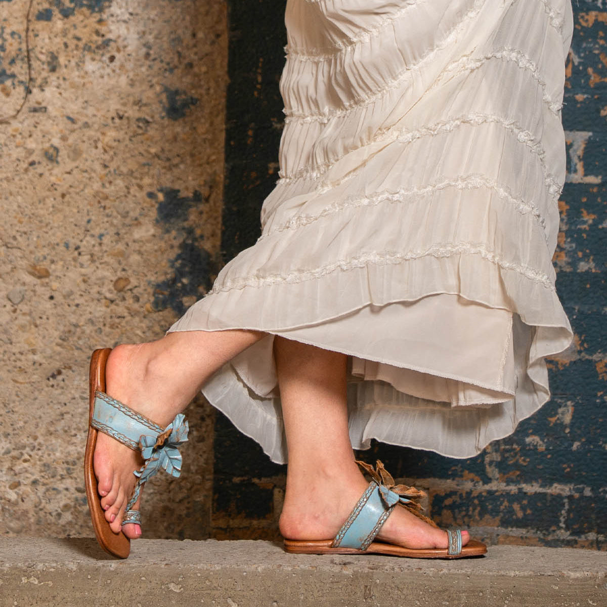 Wearing the Isobel braided strap leather sandals in light blue by Bed|Stü, a person stands on a concrete step in front of a textured wall, effortlessly embodying chic summer style in a long, cream-colored dress.