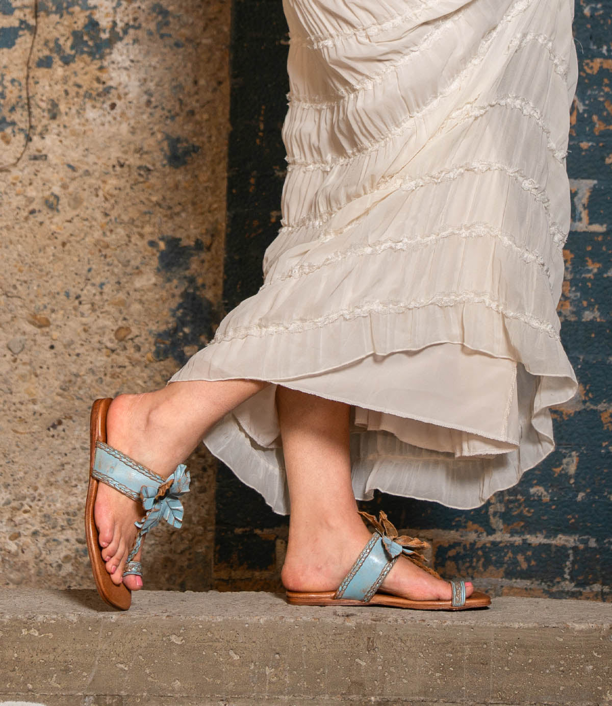 Wearing the Isobel braided strap leather sandals in light blue by Bed|Stü, a person stands on a concrete step in front of a textured wall, effortlessly embodying chic summer style in a long, cream-colored dress.