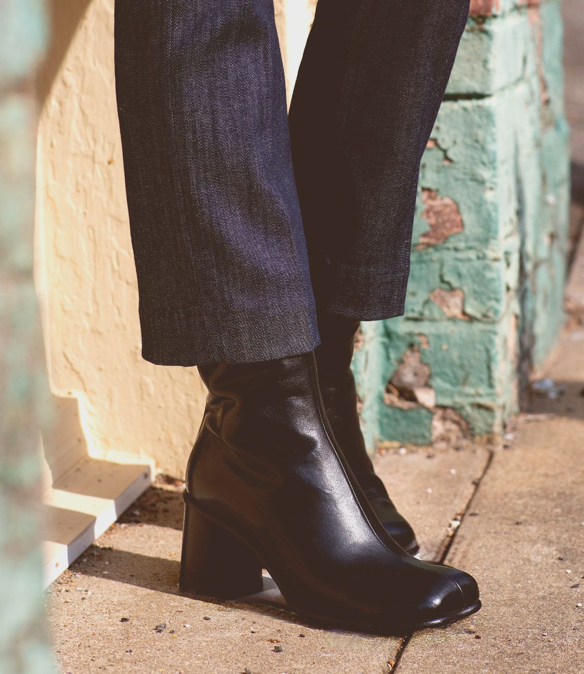 A person wearing dark pants and elegant Bed|Stü Isodora Italian leather boots with black heels stands on a sidewalk next to a textured green and yellow wall.
