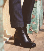 A person wearing dark pants and elegant Bed|Stü Isodora Italian leather boots with black heels stands on a sidewalk next to a textured green and yellow wall.