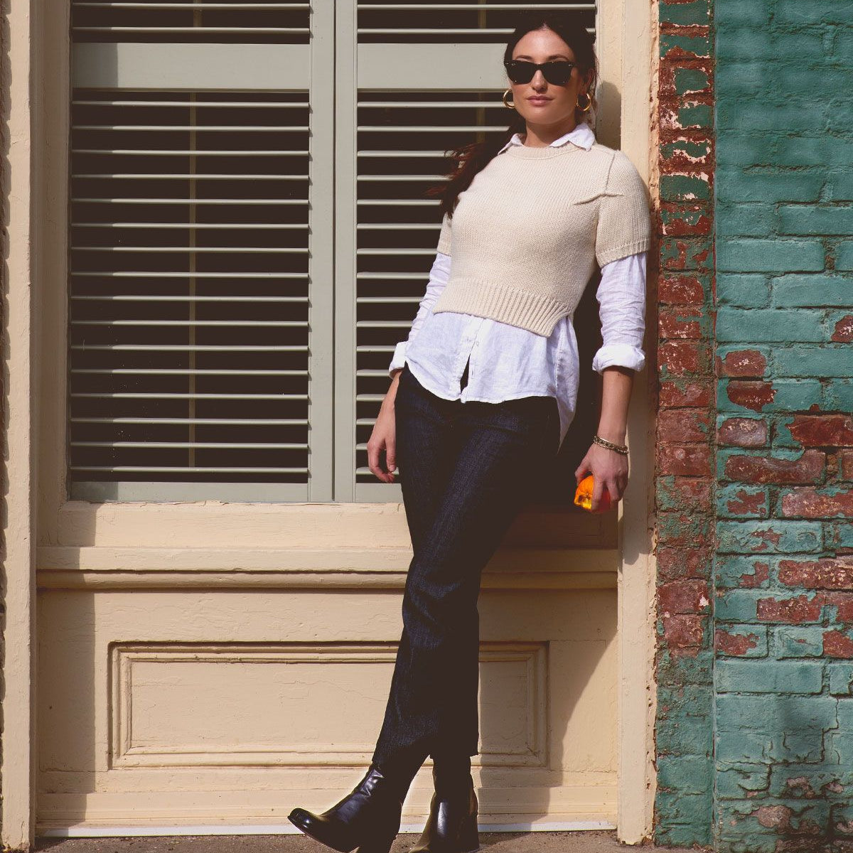 A woman in sunglasses and a white sweater, wearing the handcrafted Italian leather Bed|Stü Isodora boots, leans against a brick wall outside while holding an orange object, with closed blinds and a green-framed window in the background.