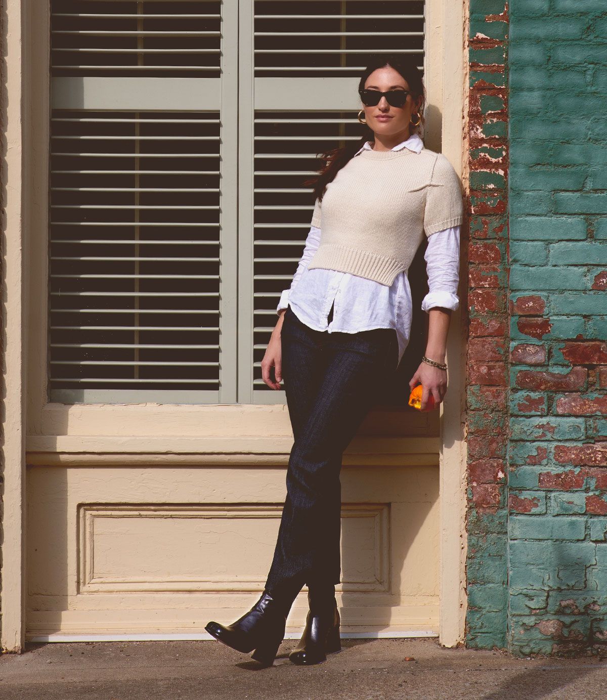 A woman in sunglasses and a white sweater, wearing the handcrafted Italian leather Bed|Stü Isodora boots, leans against a brick wall outside while holding an orange object, with closed blinds and a green-framed window in the background.