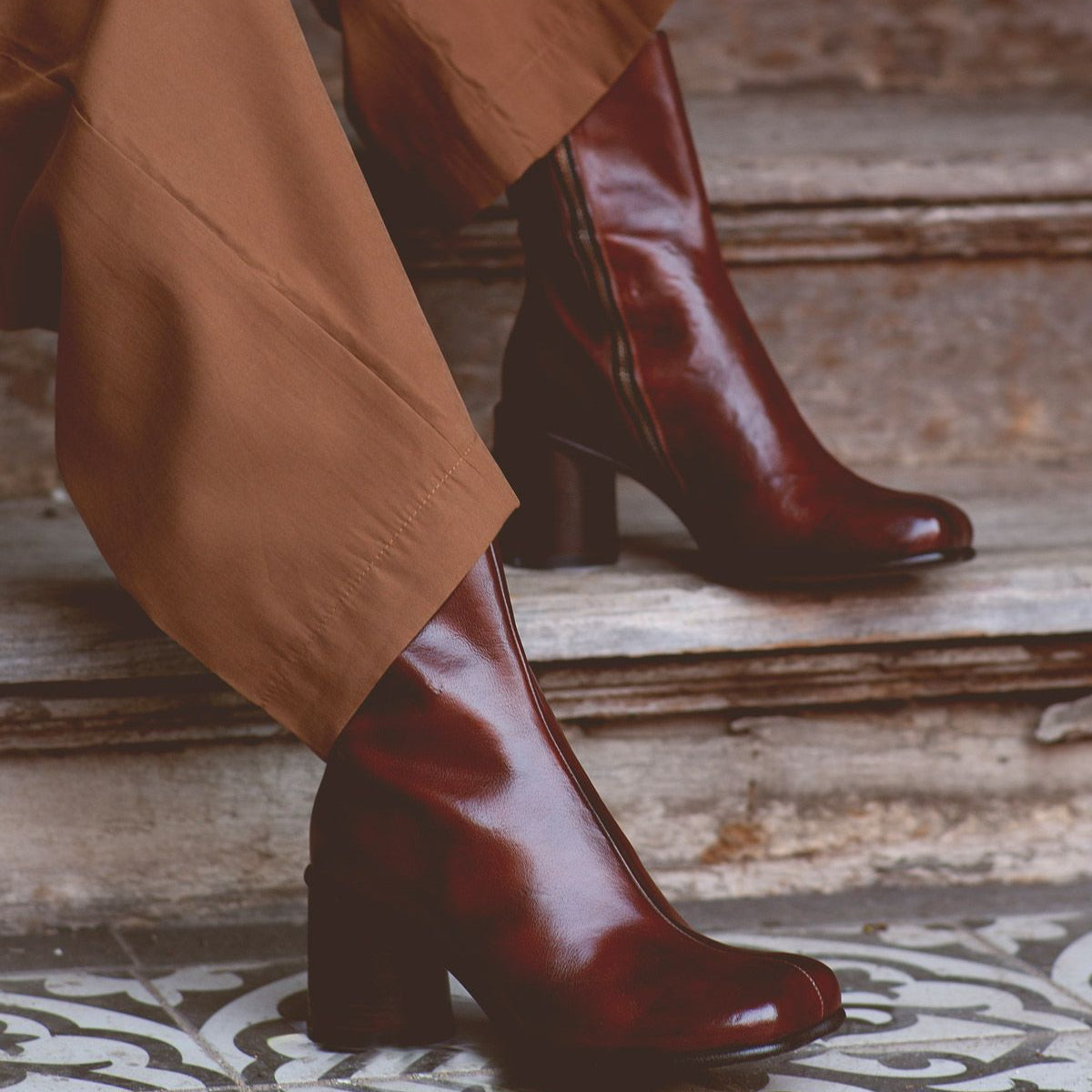 A person wearing brown pants and shiny dark brown handcrafted Bed|Stü Isodora boots stands on patterned tiles near worn stone steps.