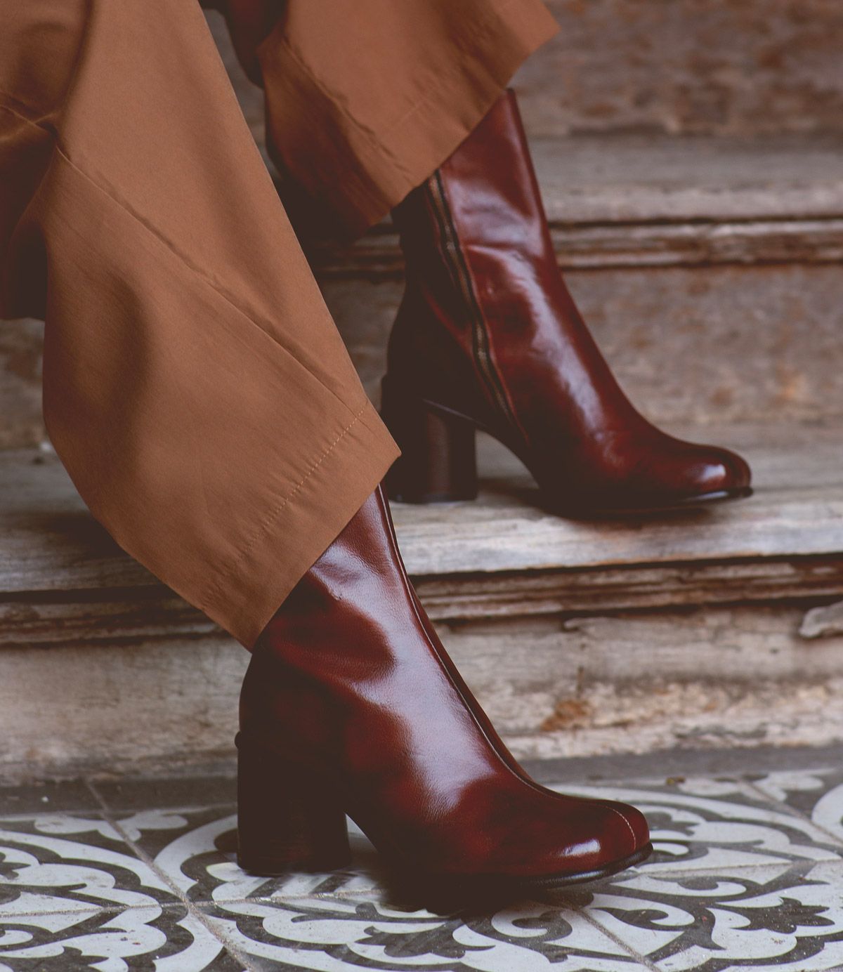 A person wearing brown pants and shiny dark brown handcrafted Bed|Stü Isodora boots stands on patterned tiles near worn stone steps.