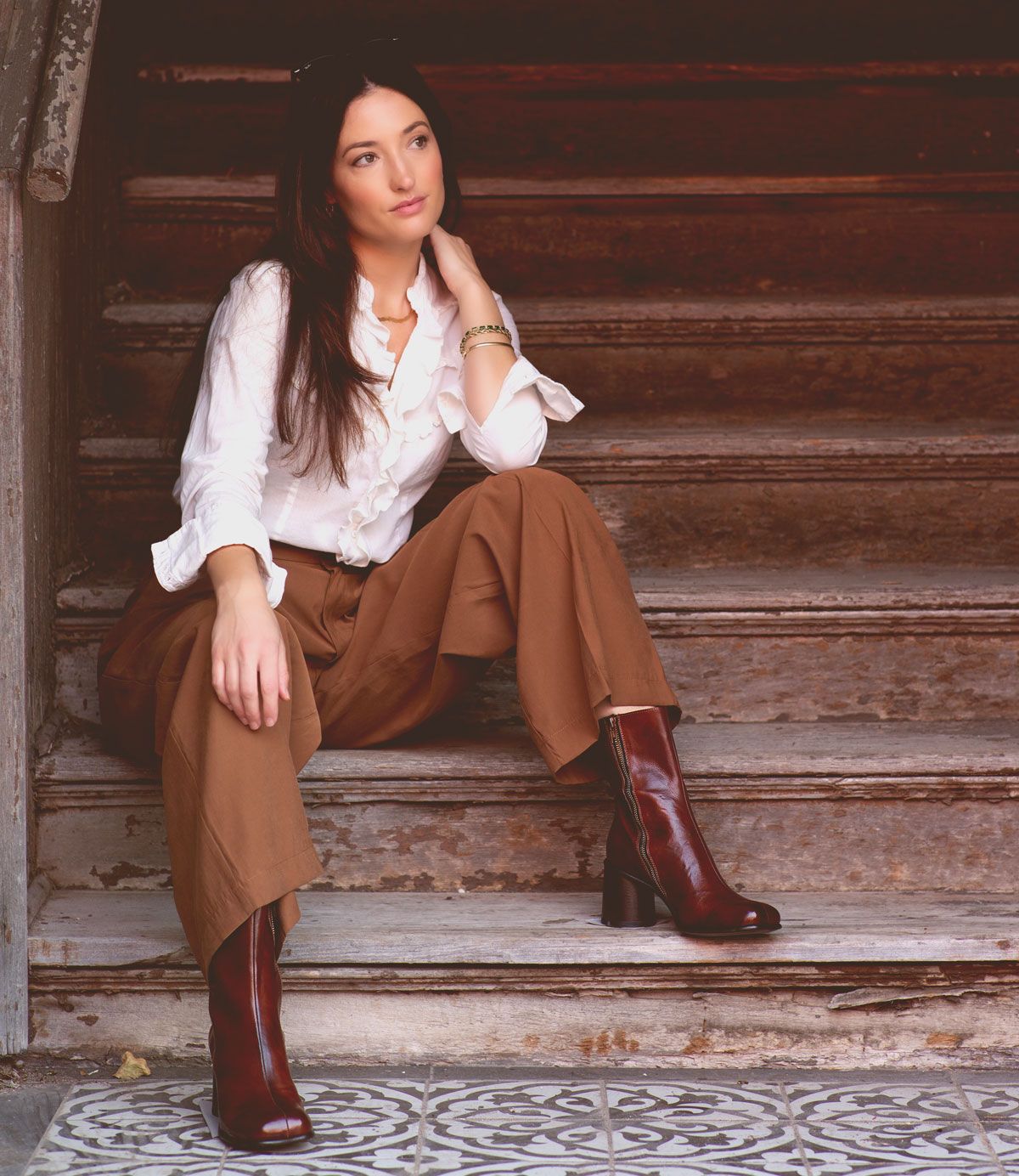 Wearing the Isodora handcrafted Italian leather boots by Bed|Stü, a woman in a white blouse and brown wide-leg pants sits on worn wooden stairs, gazing off to the side with one hand resting on her neck.